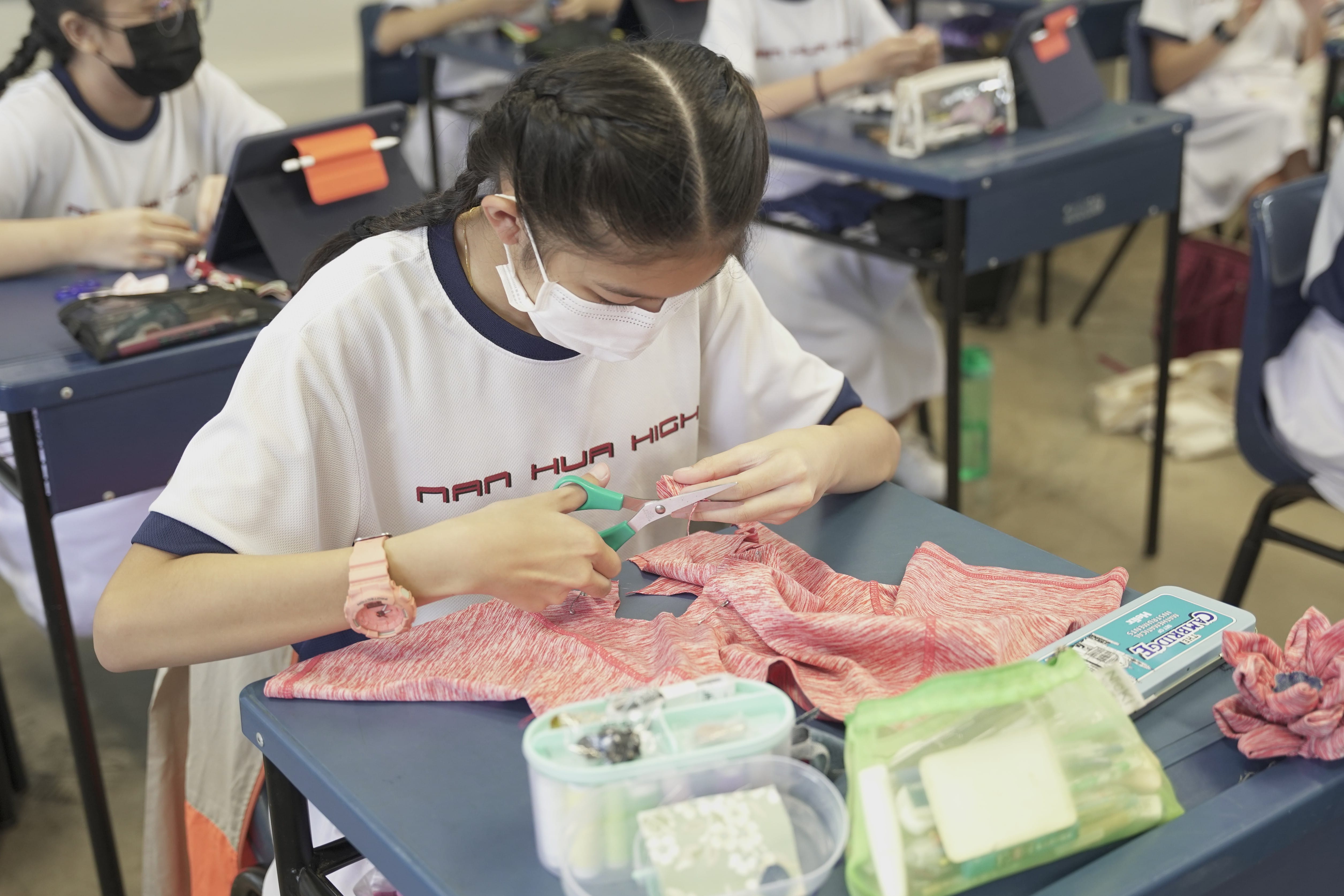 A masked schoolgirl cuts pink fabric with scissors at a desk in a classroom with other students nearby.